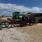 Wood processor attached to a green tractor in a gravel yard, with stacked logs and a shed in the background.