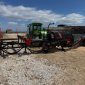Green tractor attached to red-and-black roadside wood processor on a rocky site with stacked logs in the background.