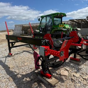 1000047113 Red and black Palax MIDI wood splitter/processor attached to a trailer, with a green tractor in a lumber yard setting.