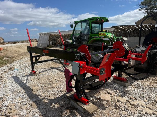 1000047113 Red and black Palax MIDI wood splitter/processor attached to a trailer, with a green tractor in a lumber yard setting.