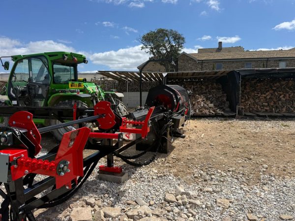 1000047114 Green tractor with attached red farming equipment on a gravel yard; woodpile and stone house in background.