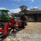 Green tractor with attached red farming equipment on a gravel yard; woodpile and stone house in background.